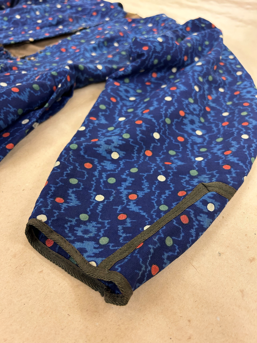 Klänning i blå bottenfärg, vattrat mönster, prickar i gult, rött och grönt. Slätt figurskuret liv med insydda fjädrar. Valben i ryggsöm och insnitt fram. Knäppning framtill med 13 st hyskor och hakar, varav flera saknas. Ingen krage. Isydd ärm med lagda veck över axeln. Passpoal i sömmen. Smalt markerat ryggstycke med svarta passpoalmarkerade sömmar. En vid ärm som smalnar vid handleden, vilken är kantad med svarta bomullsband, vilka nu är gröna. Kjolen har lagda veck runt om, sprund mitt fram. Sydd av 4 hela våder och en kilskuren våd. Sparkband av samma bruna linnelärft som livets foder. Vänster ärm är fodrad med brunt kattuntyg med mönster av vita prickar på brun botten, däröver brandgula blommor och streckliknande mönster. Klänningen är lagad på flera ställen och hålig.