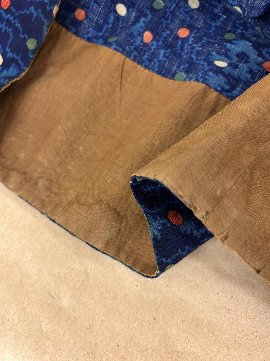 Klänning i blå bottenfärg, vattrat mönster, prickar i gult, rött och grönt. Slätt figurskuret liv med insydda fjädrar. Valben i ryggsöm och insnitt fram. Knäppning framtill med 13 st hyskor och hakar, varav flera saknas. Ingen krage. Isydd ärm med lagda veck över axeln. Passpoal i sömmen. Smalt markerat ryggstycke med svarta passpoalmarkerade sömmar. En vid ärm som smalnar vid handleden, vilken är kantad med svarta bomullsband, vilka nu är gröna. Kjolen har lagda veck runt om, sprund mitt fram. Sydd av 4 hela våder och en kilskuren våd. Sparkband av samma bruna linnelärft som livets foder. Vänster ärm är fodrad med brunt kattuntyg med mönster av vita prickar på brun botten, däröver brandgula blommor och streckliknande mönster. Klänningen är lagad på flera ställen och hålig.