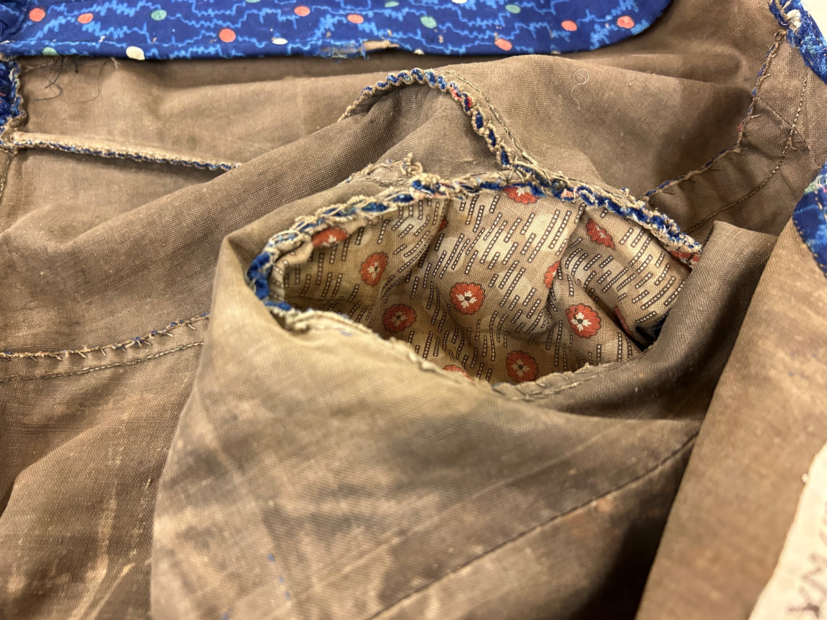 Klänning i blå bottenfärg, vattrat mönster, prickar i gult, rött och grönt. Slätt figurskuret liv med insydda fjädrar. Valben i ryggsöm och insnitt fram. Knäppning framtill med 13 st hyskor och hakar, varav flera saknas. Ingen krage. Isydd ärm med lagda veck över axeln. Passpoal i sömmen. Smalt markerat ryggstycke med svarta passpoalmarkerade sömmar. En vid ärm som smalnar vid handleden, vilken är kantad med svarta bomullsband, vilka nu är gröna. Kjolen har lagda veck runt om, sprund mitt fram. Sydd av 4 hela våder och en kilskuren våd. Sparkband av samma bruna linnelärft som livets foder. Vänster ärm är fodrad med brunt kattuntyg med mönster av vita prickar på brun botten, däröver brandgula blommor och streckliknande mönster. Klänningen är lagad på flera ställen och hålig.