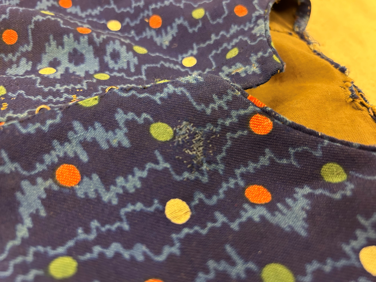 Klänning i blå bottenfärg, vattrat mönster, prickar i gult, rött och grönt. Slätt figurskuret liv med insydda fjädrar. Valben i ryggsöm och insnitt fram. Knäppning framtill med 13 st hyskor och hakar, varav flera saknas. Ingen krage. Isydd ärm med lagda veck över axeln. Passpoal i sömmen. Smalt markerat ryggstycke med svarta passpoalmarkerade sömmar. En vid ärm som smalnar vid handleden, vilken är kantad med svarta bomullsband, vilka nu är gröna. Kjolen har lagda veck runt om, sprund mitt fram. Sydd av 4 hela våder och en kilskuren våd. Sparkband av samma bruna linnelärft som livets foder. Vänster ärm är fodrad med brunt kattuntyg med mönster av vita prickar på brun botten, däröver brandgula blommor och streckliknande mönster. Klänningen är lagad på flera ställen och hålig.