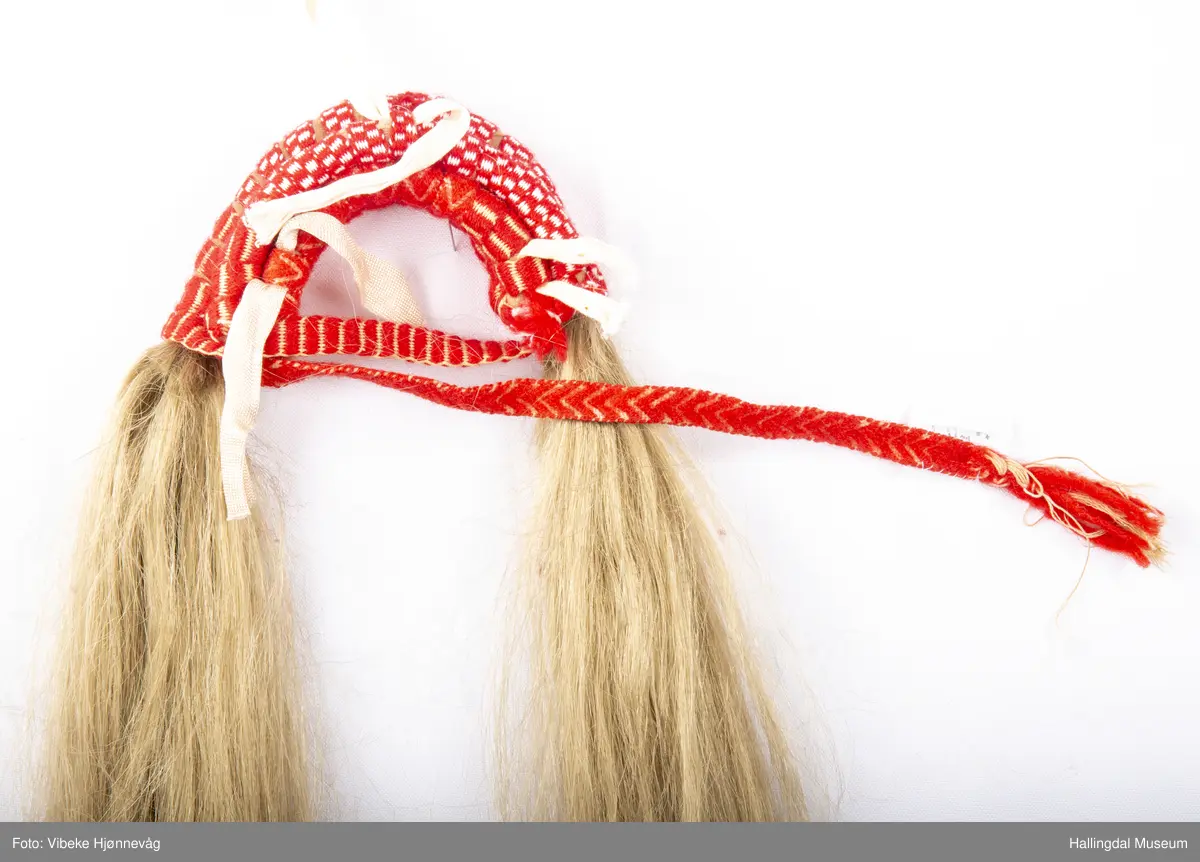 Flette med hår til hodebunad fra Øvre Hallingdal. Båndet er lagd av 3 ulike bånd; klauveband, kambeband og et annet vevd ullbånd. Håret er ca. 40 cm langt. Ull- og bomullsgarn vevd på grindvev. Rødt og hvitt.