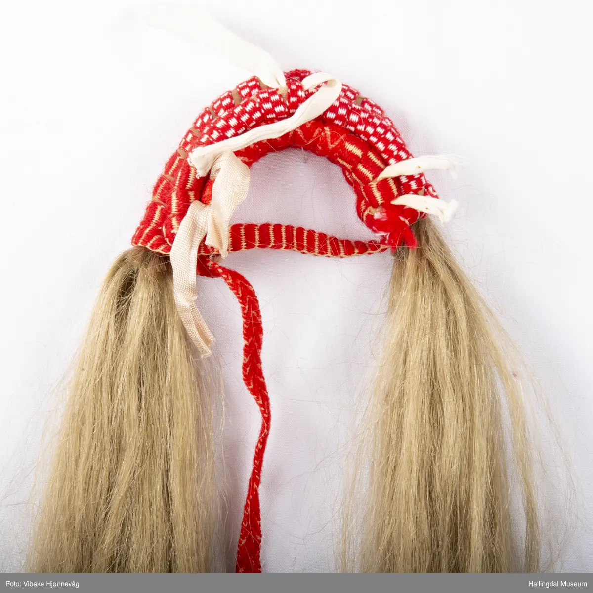 Flette med hår til hodebunad fra Øvre Hallingdal. Båndet er lagd av 3 ulike bånd; klauveband, kambeband og et annet vevd ullbånd. Håret er ca. 40 cm langt. Ull- og bomullsgarn vevd på grindvev. Rødt og hvitt.