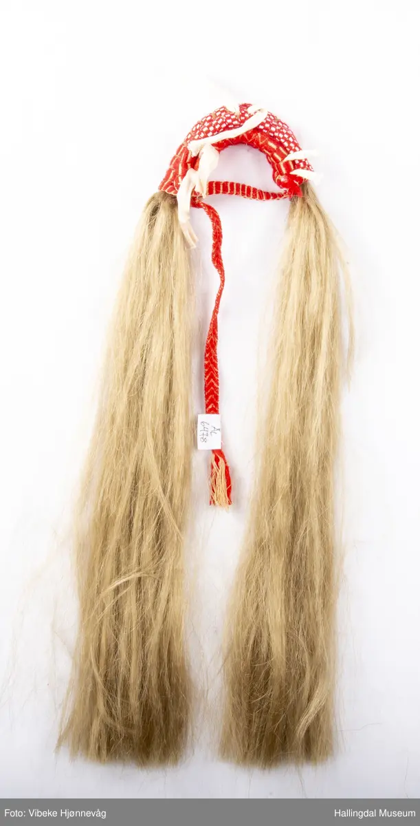Flette med hår til hodebunad fra Øvre Hallingdal. Båndet er lagd av 3 ulike bånd; klauveband, kambeband og et annet vevd ullbånd. Håret er ca. 40 cm langt. Ull- og bomullsgarn vevd på grindvev. Rødt og hvitt.