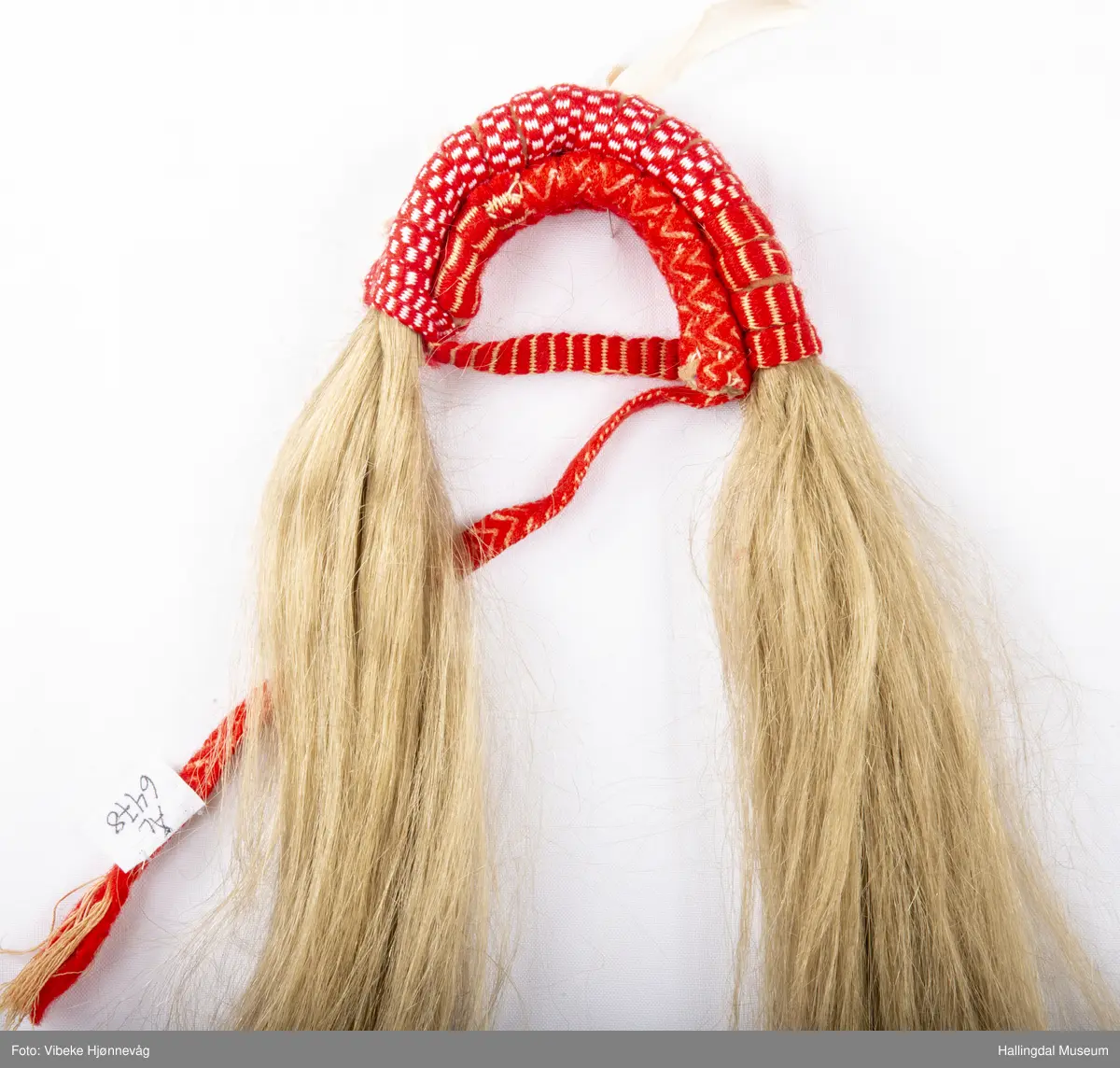 Flette med hår til hodebunad fra Øvre Hallingdal. Båndet er lagd av 3 ulike bånd; klauveband, kambeband og et annet vevd ullbånd. Håret er ca. 40 cm langt. Ull- og bomullsgarn vevd på grindvev. Rødt og hvitt.