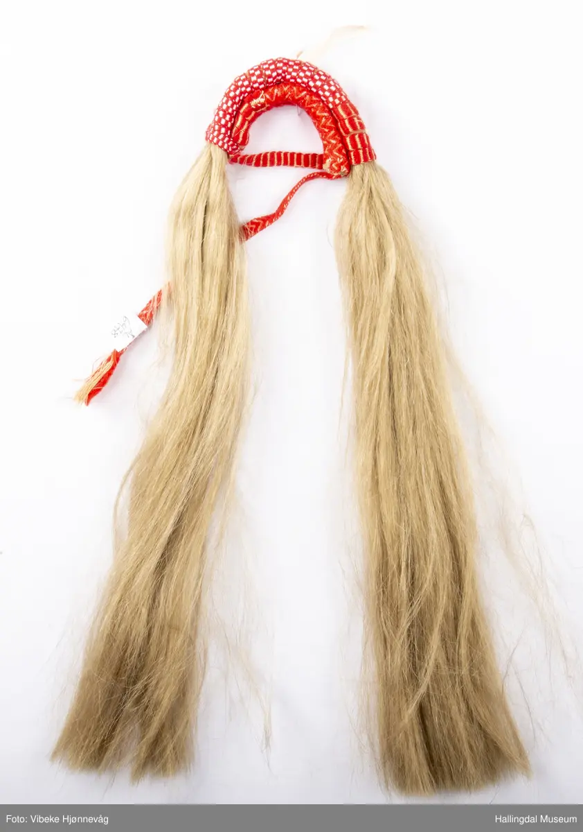 Flette med hår til hodebunad fra Øvre Hallingdal. Båndet er lagd av 3 ulike bånd; klauveband, kambeband og et annet vevd ullbånd. Håret er ca. 40 cm langt. Ull- og bomullsgarn vevd på grindvev. Rødt og hvitt.