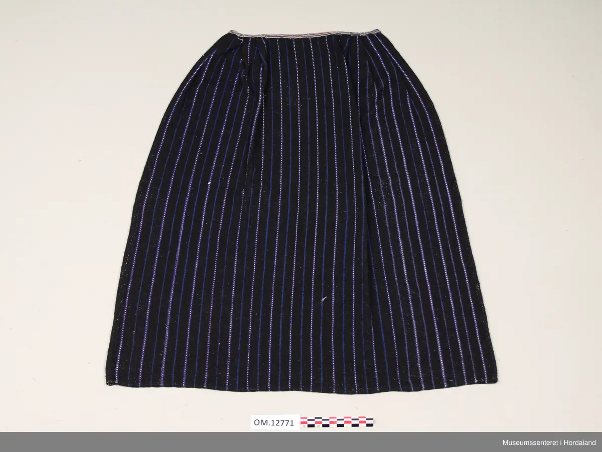 Heimevove ullforkle i mørk blått med kvitt og mellomblått mønster - mønsteret er på langs av forkleet. Det kvite er bomullsgarn. Oppe i livet er det sydd på eit kvitt og gråstripete bomullsstoff og under dette bandet ligg forkleet i folder. I begge endane på bandet er det ei metallhekte til feste av forkleband.