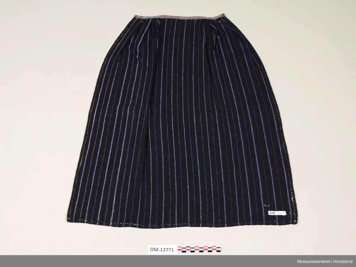 Heimevove ullforkle i mørk blått med kvitt og mellomblått mønster - mønsteret er på langs av forkleet. Det kvite er bomullsgarn. Oppe i livet er det sydd på eit kvitt og gråstripete bomullsstoff og under dette bandet ligg forkleet i folder. I begge endane på bandet er det ei metallhekte til feste av forkleband.