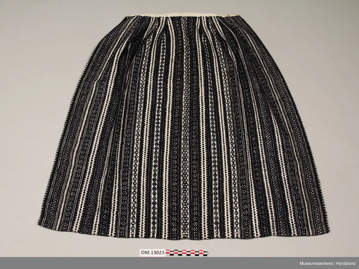 Mørkeblått vove ullforkle med mange ulike mønsterstriper i kvitt. Skoning på oppleggskanten i mørkeblått stoff med prikkar. Fire folder på kvar side av slett midtparti. Kvitt band til linning. Ringar i kvar side av linninga.