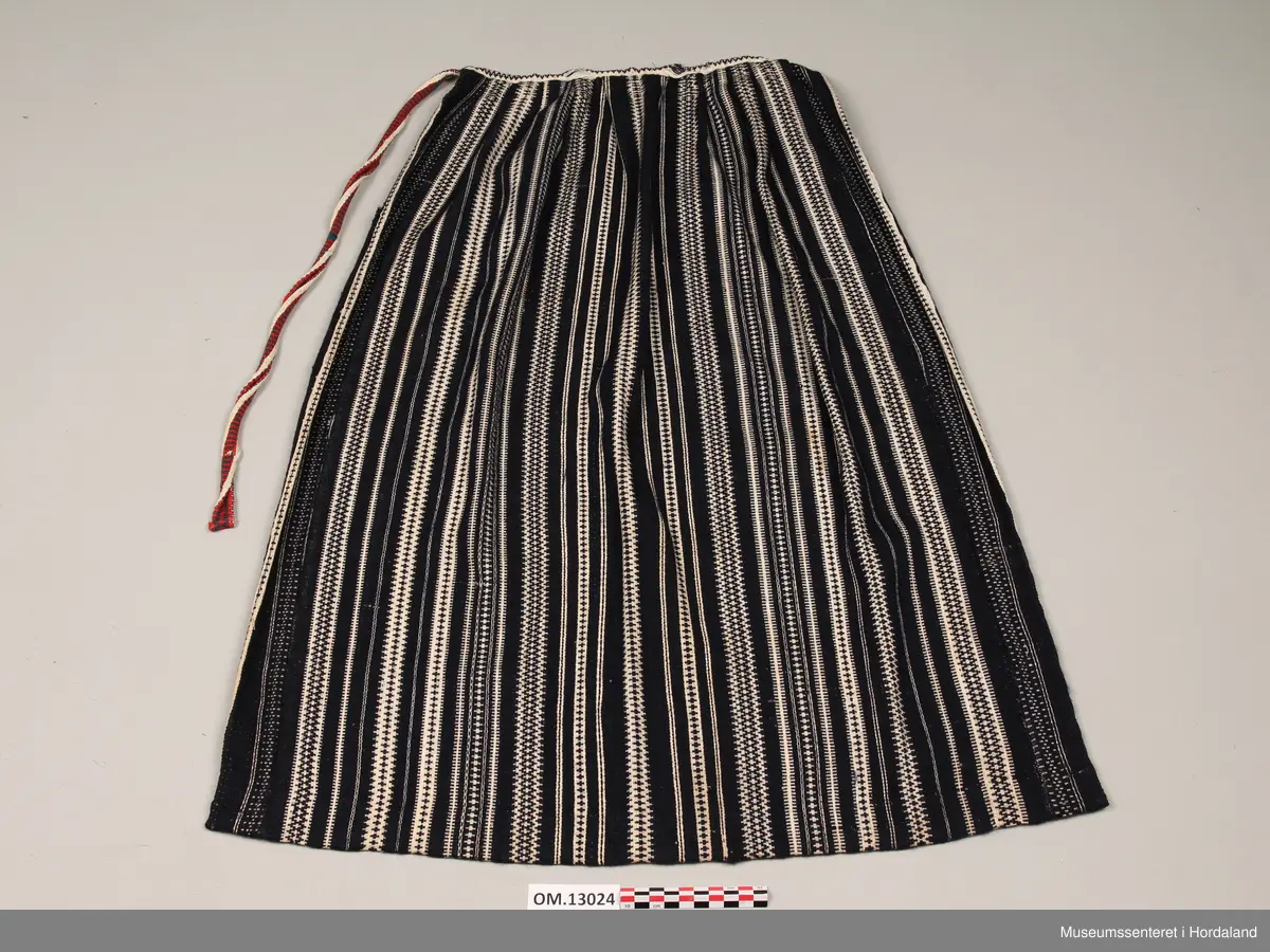 Mørkeblått vove ullforkle med langsgåande mønsterstriper i kvitt. Kvit skoning på oppleggskanten. Breidda av forkleet er auka på kvar side med ei stoffstrimle med ei bord ulik dei andre bordene. Fem folder på kvar side av eit smalt slett midtparti. Vove band til linning i same fargane som resten av forkleet. Voven beltetamp sydd på i høgre side i fargane kvitt, raudt og grønt.
