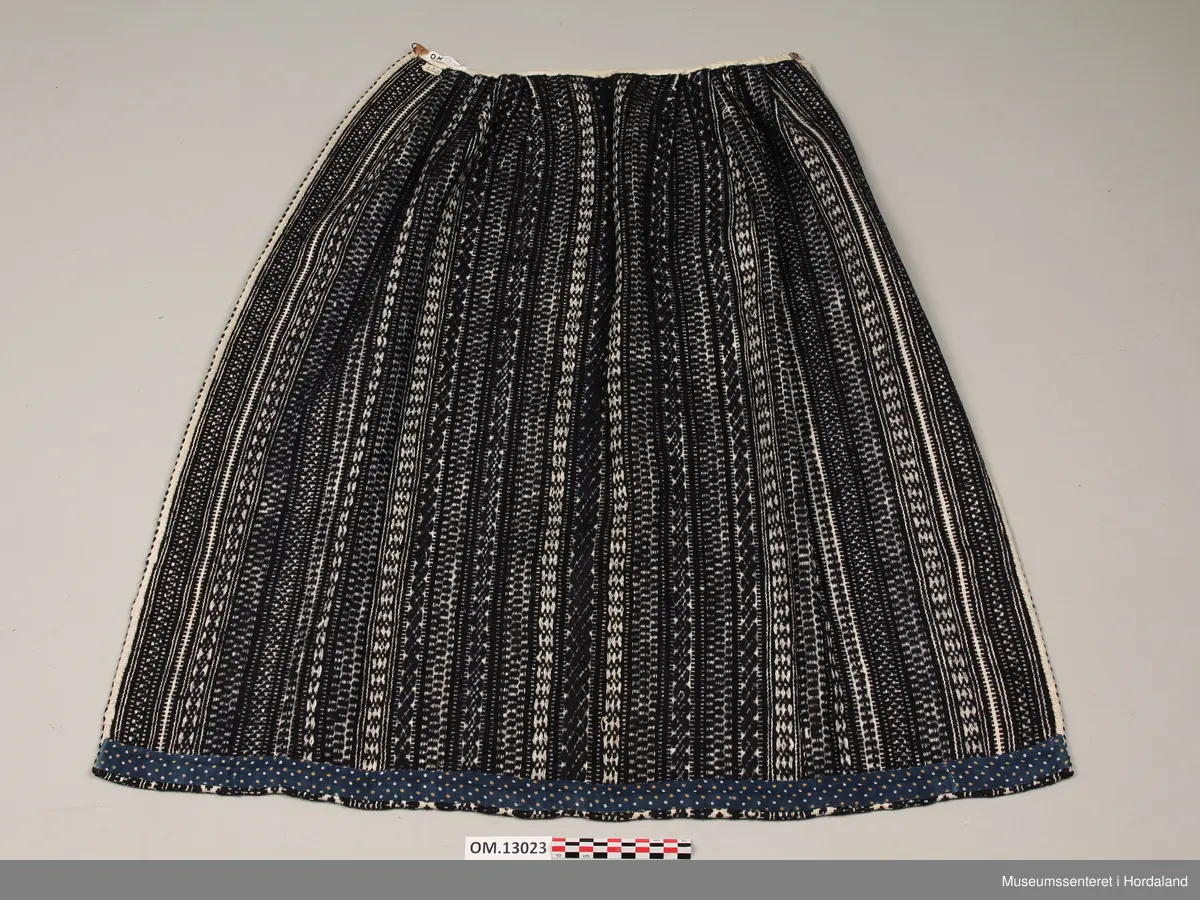 Mørkeblått vove ullforkle med mange ulike mønsterstriper i kvitt. Skoning på oppleggskanten i mørkeblått stoff med prikkar. Fire folder på kvar side av slett midtparti. Kvitt band til linning. Ringar i kvar side av linninga.