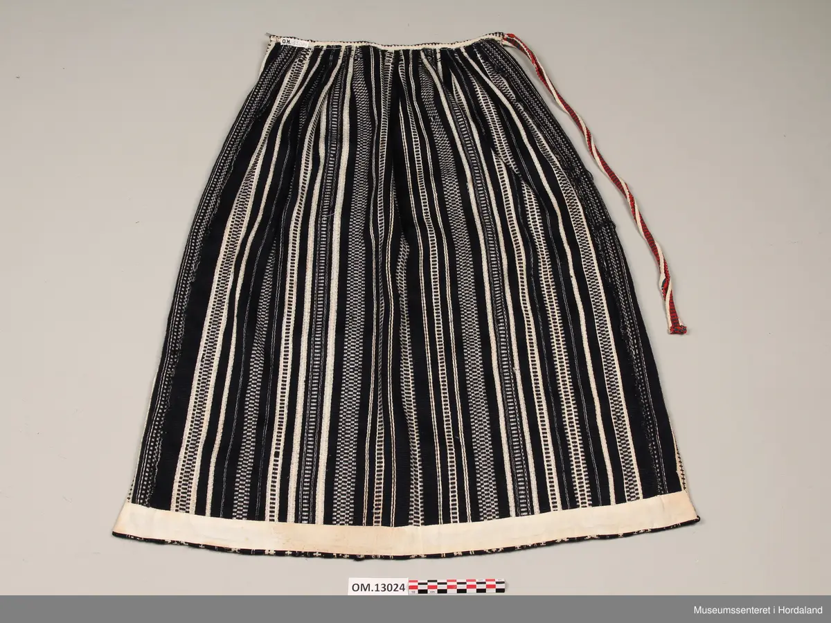 Mørkeblått vove ullforkle med langsgåande mønsterstriper i kvitt. Kvit skoning på oppleggskanten. Breidda av forkleet er auka på kvar side med ei stoffstrimle med ei bord ulik dei andre bordene. Fem folder på kvar side av eit smalt slett midtparti. Vove band til linning i same fargane som resten av forkleet. Voven beltetamp sydd på i høgre side i fargane kvitt, raudt og grønt.