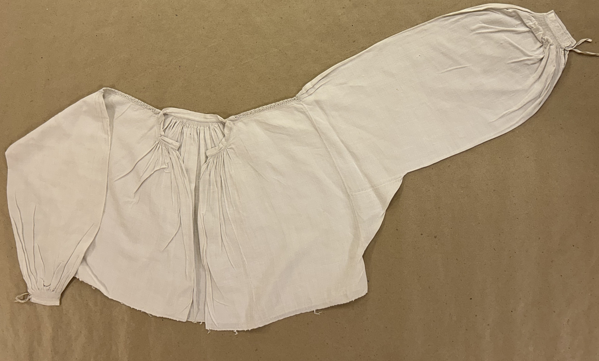 Skjortöverdel, "opplott", av handvävt hellinne. Smal halslinning, fina rynkor mitt bak och närmast sprundet fram. Från halslinning till ärmsöm en knypplad spets. På ärmen rynkor endast mitt för spetsen. Smal handlinning, broderad i vitt. Knytes med smala, handvävda linneband. Brett rynkparti.