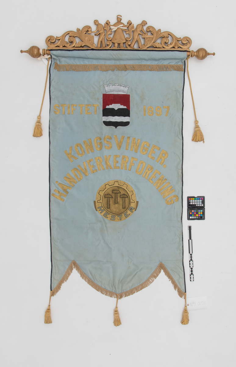 Faneduk: Byvåpen Kongsvinger kommune. Symbol for håndverkerforeningen. 
Toppstykke: I midten en baker, til høyre en snekker og til venstre en murer. Akantusranker på begge sider av disse. Tosidig, samme motiv.