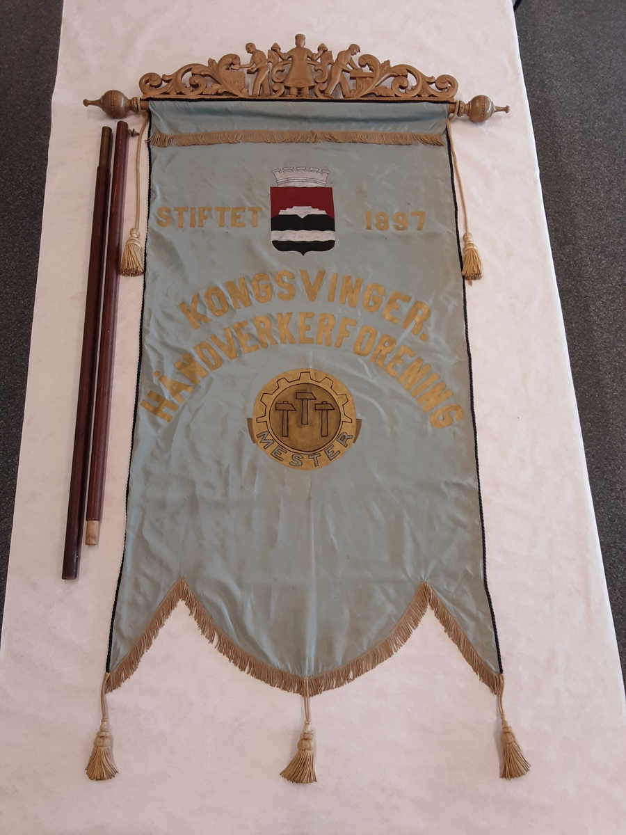 Faneduk: Byvåpen Kongsvinger kommune. Symbol for håndverkerforeningen. 
Toppstykke: I midten en baker, til høyre en snekker og til venstre en murer. Akantusranker på begge sider av disse. Tosidig, samme motiv.