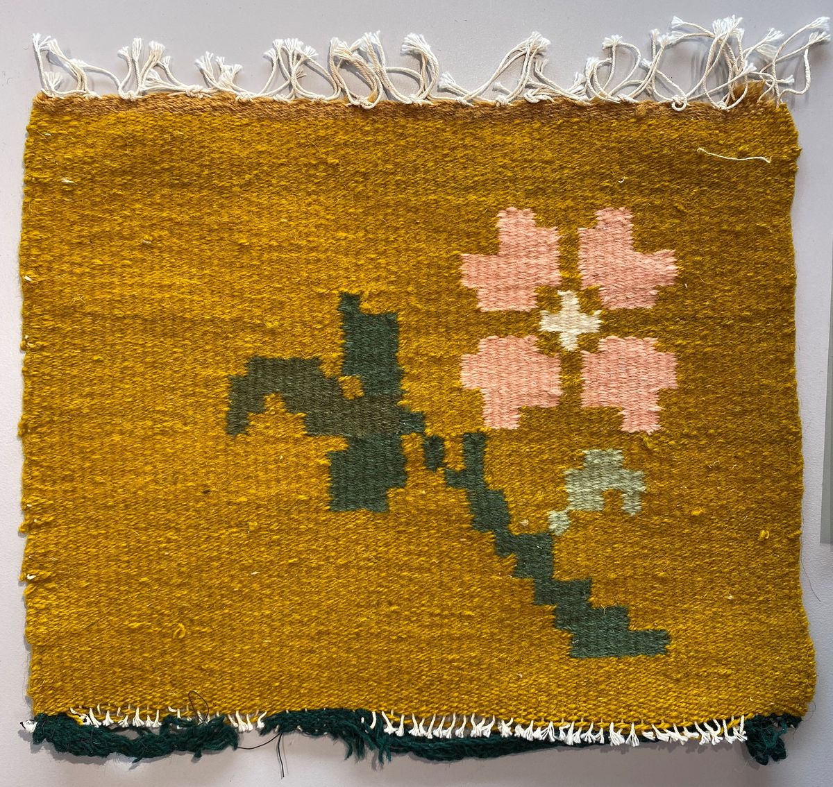 Vevd bilde av en rosa blomst på sennepsgul bakgrunn. Veven er avsluttet med en grønn kant som har delvis løsnet.
