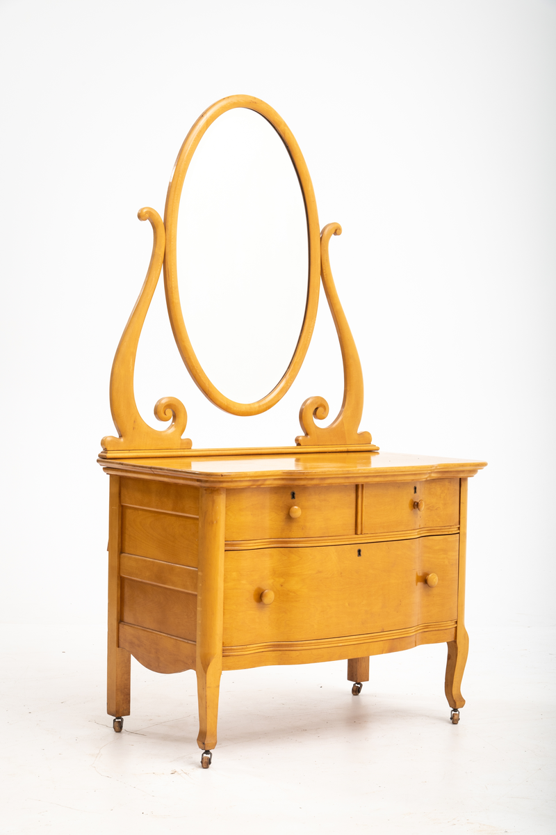 Kommode. Topplate og fronten på kommoden er bølget i formen. To mindre skuffer øverst, og en stor låsbar skuff nederst. Treknotter. Ben med hjul.
