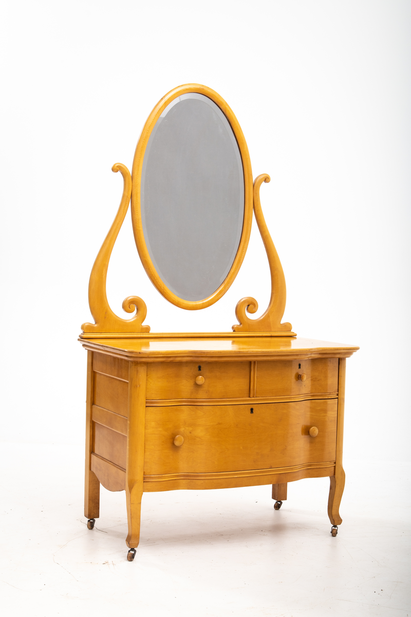 Kommode. Topplate og fronten på kommoden er bølget i formen. To mindre skuffer øverst, og en stor låsbar skuff nederst. Treknotter. Ben med hjul.