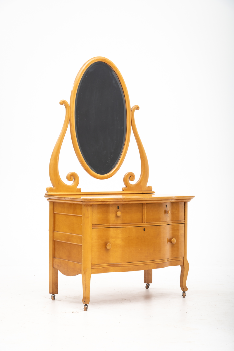 Kommode. Topplate og fronten på kommoden er bølget i formen. To mindre skuffer øverst, og en stor låsbar skuff nederst. Treknotter. Ben med hjul.