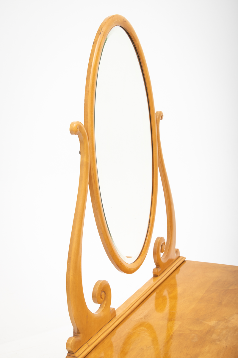 Ovalt speil i ramme. Speilet er festet til et stativ i hver side. Sidestykkene på stativet har S-form og utskjæringer og er festet til en fotlist. Denne fotlisten festes på toppen av kommoden.