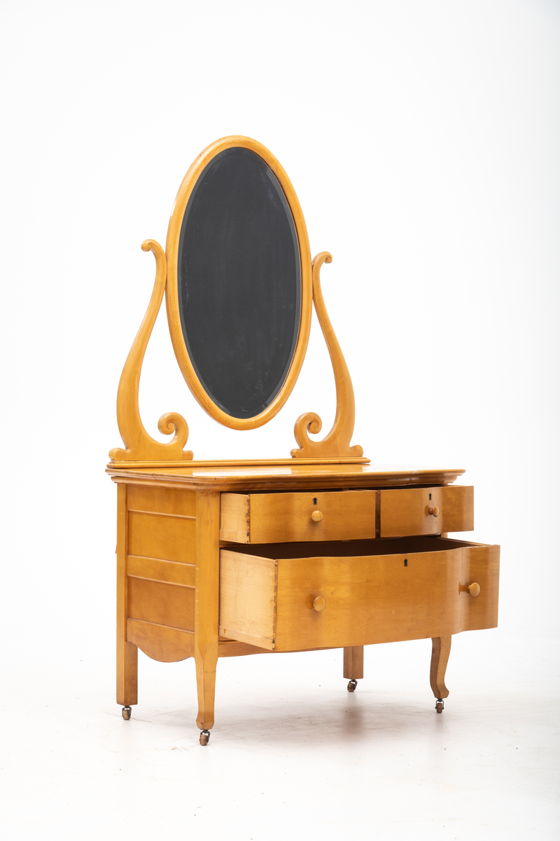 Kommode. Topplate og fronten på kommoden er bølget i formen. To mindre skuffer øverst, og en stor låsbar skuff nederst. Treknotter. Ben med hjul.