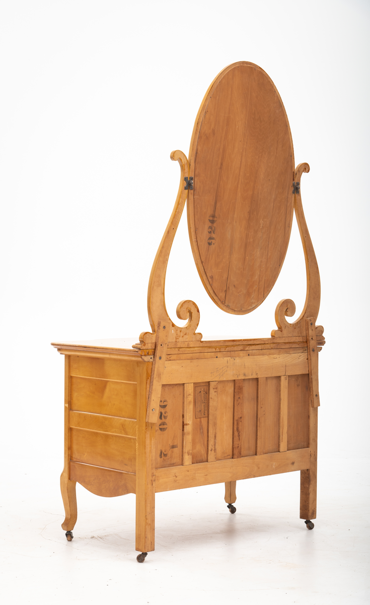 Ovalt speil i ramme. Speilet er festet til et stativ i hver side. Sidestykkene på stativet har S-form og utskjæringer og er festet til en fotlist. Denne fotlisten festes på toppen av kommoden.