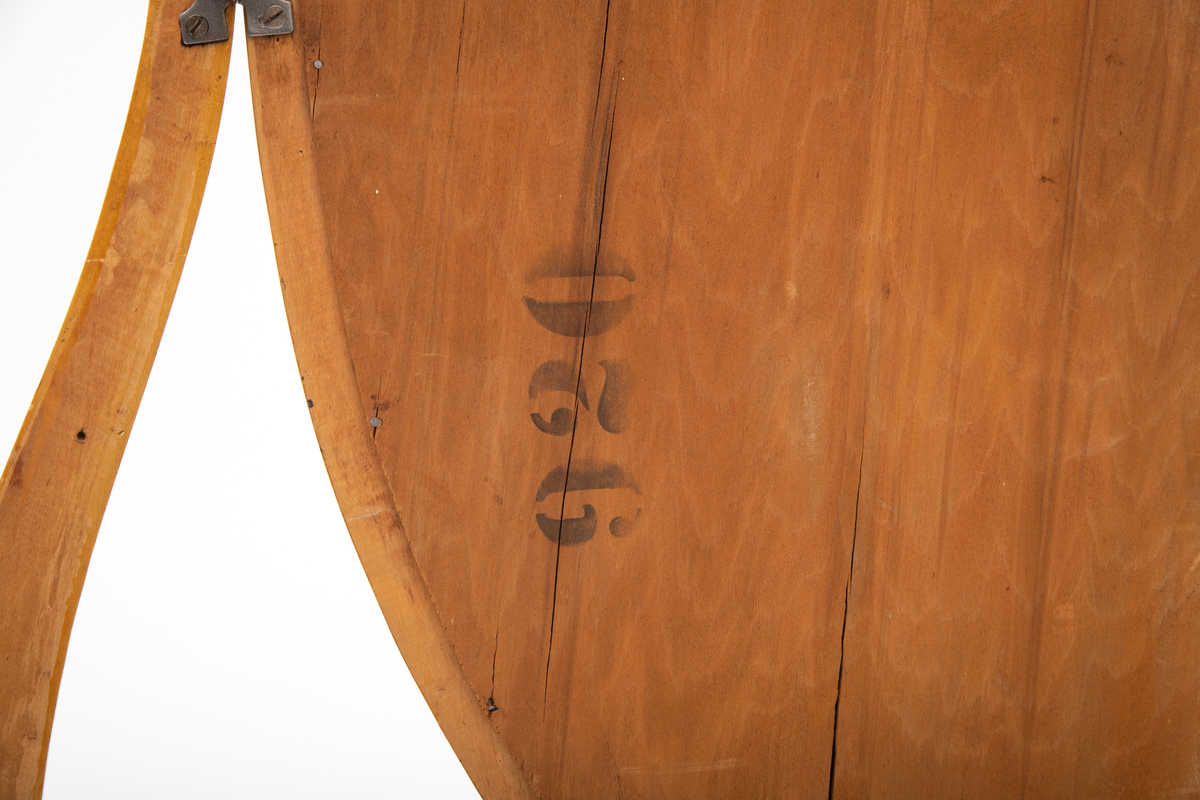 Ovalt speil i ramme. Speilet er festet til et stativ i hver side. Sidestykkene på stativet har S-form og utskjæringer og er festet til en fotlist. Denne fotlisten festes på toppen av kommoden.