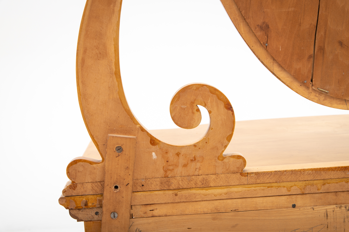 Ovalt speil i ramme. Speilet er festet til et stativ i hver side. Sidestykkene på stativet har S-form og utskjæringer og er festet til en fotlist. Denne fotlisten festes på toppen av kommoden.