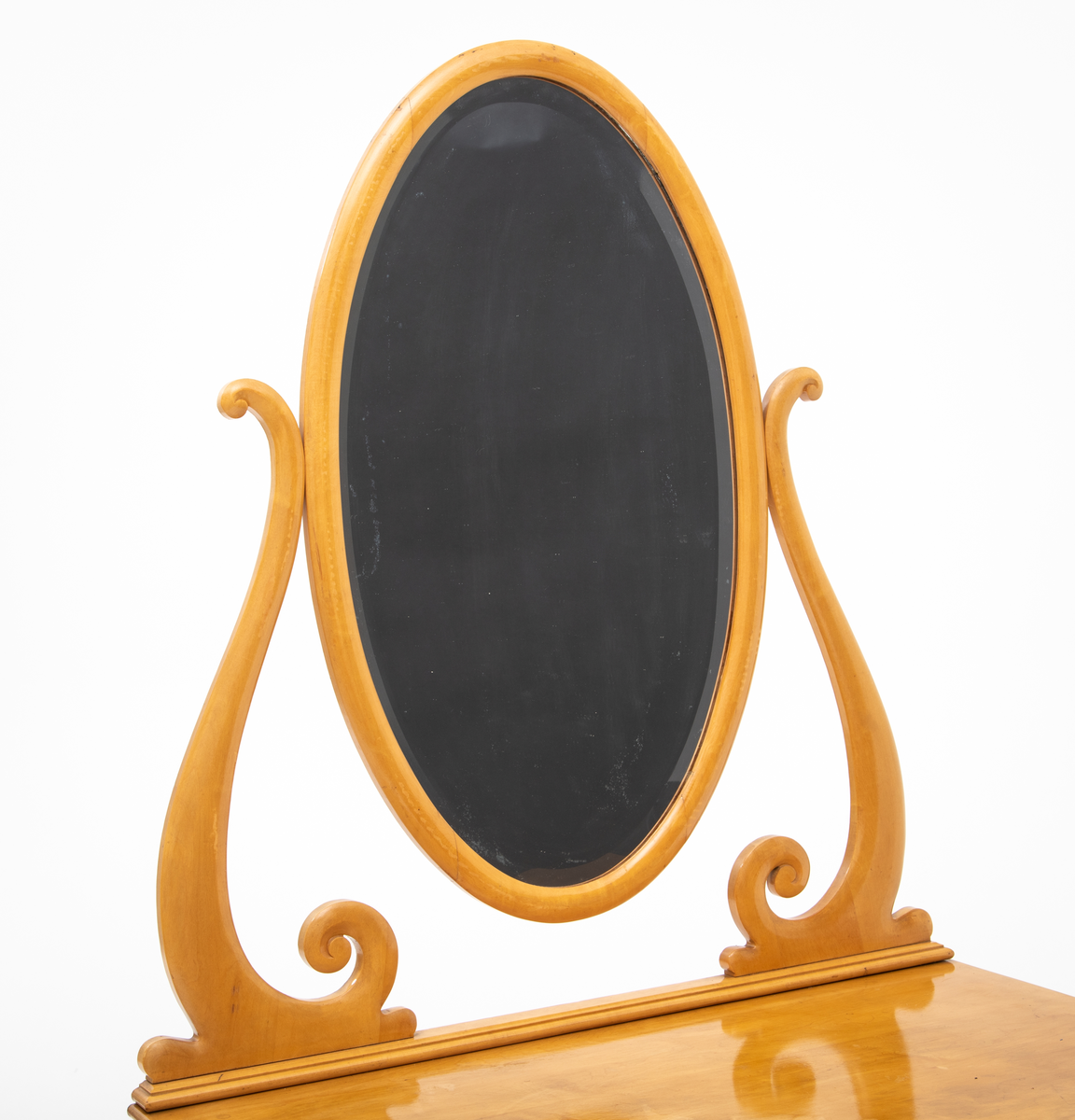 Ovalt speil i ramme. Speilet er festet til et stativ i hver side. Sidestykkene på stativet har S-form og utskjæringer og er festet til en fotlist. Denne fotlisten festes på toppen av kommoden.