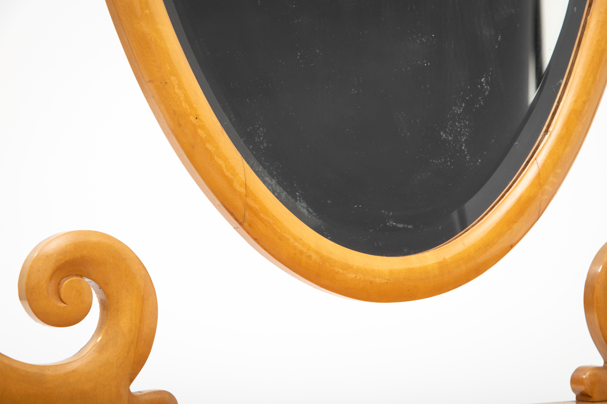 Ovalt speil i ramme. Speilet er festet til et stativ i hver side. Sidestykkene på stativet har S-form og utskjæringer og er festet til en fotlist. Denne fotlisten festes på toppen av kommoden.