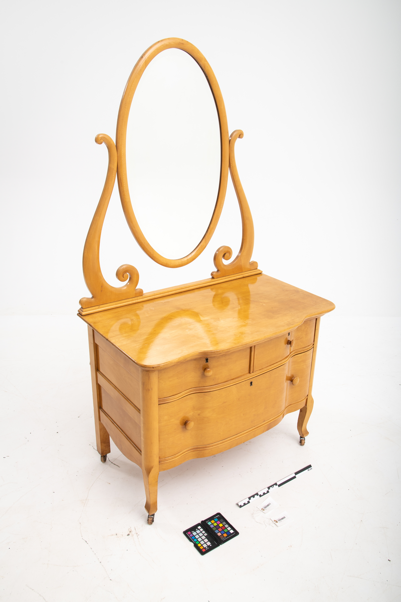 Ovalt speil i ramme. Speilet er festet til et stativ i hver side. Sidestykkene på stativet har S-form og utskjæringer og er festet til en fotlist. Denne fotlisten festes på toppen av kommoden.