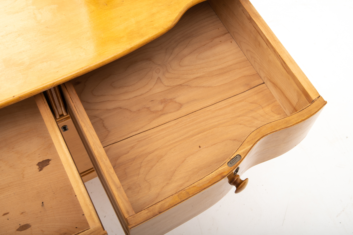 Kommode. Topplate og fronten på kommoden er bølget i formen. To mindre skuffer øverst, og en stor låsbar skuff nederst. Treknotter. Ben med hjul.