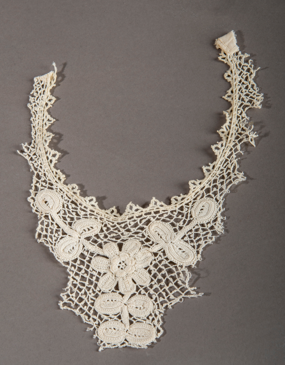 Blondekrage/kjolepynt. Ekryfarget. Motiv midt foran av en blomst med stilk og blader.