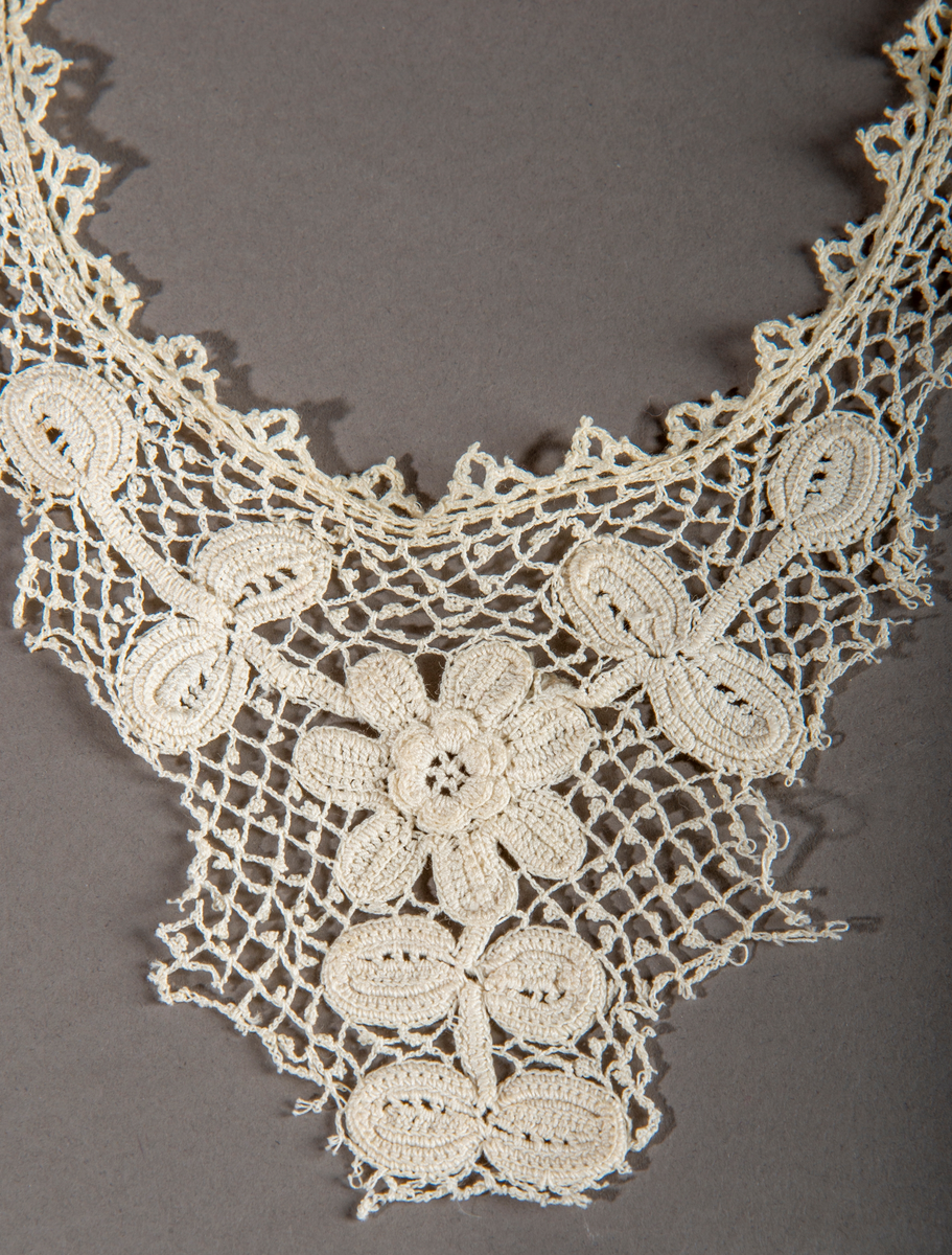 Blondekrage/kjolepynt. Ekryfarget. Motiv midt foran av en blomst med stilk og blader.