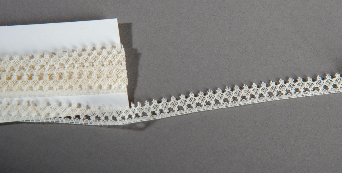 Blonderemse. Hvit. Smal. "Hullkant" langs den ene siden, taggete langs den andre.