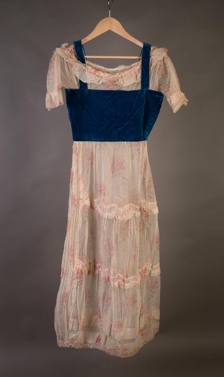 Kjole med fløyelsliv med stropper. Tyllermer, kanting over livet samt skjørtet. Skjørt med bisesømmer og rynkefelt. Lukning i siden med hekter.