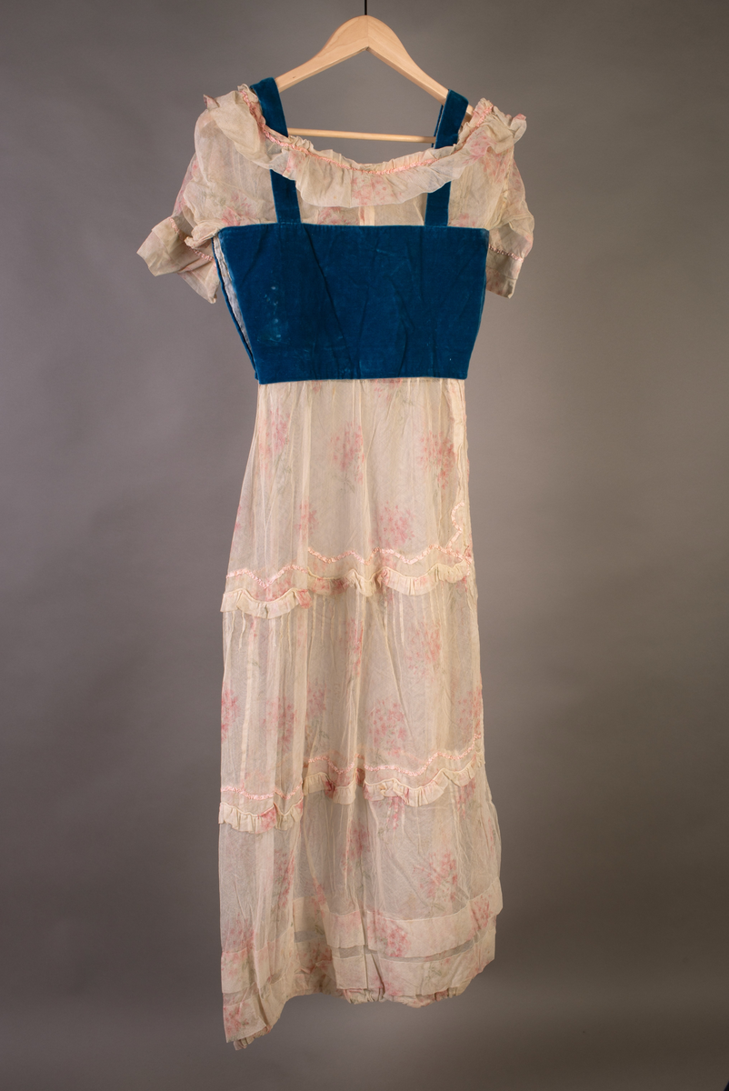 Kjole med fløyelsliv med stropper. Tyllermer, kanting over livet samt skjørtet. Skjørt med bisesømmer og rynkefelt. Lukning i siden med hekter.