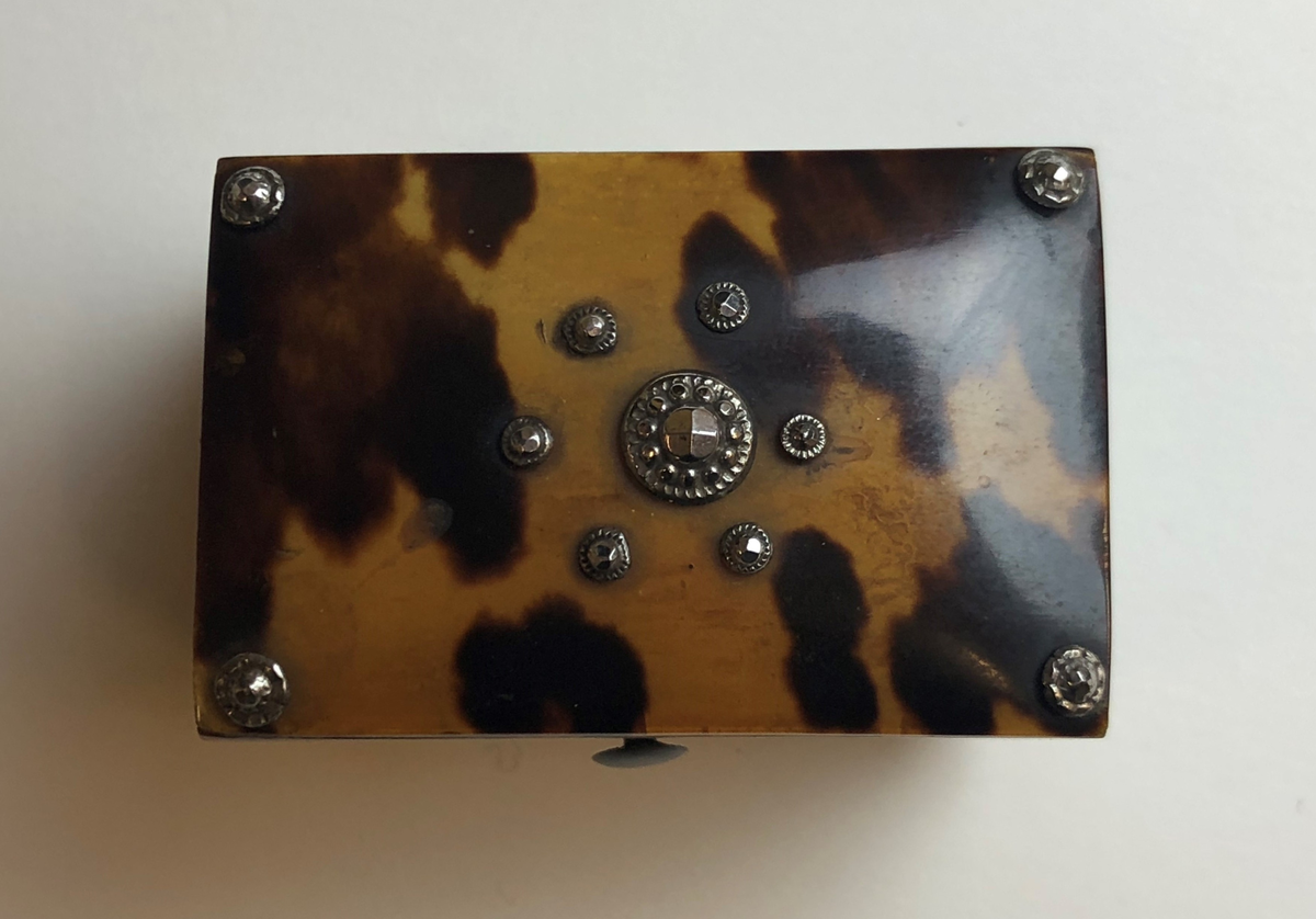 Material 4: Papper.
Rektangulär form, underdel med lock som öppnas och stängs med hjälp av ett gångjärn i metall. Av trä,  på utsidan klädd med en yta av ljusbrun plast med mörkbruna fläckar, som ska likna ädelträ. Locket något välvt vilket gör att asken liknar en kista. Underdelens insida klädd med rosa papper, lockets insida klädd med sammet. Lockets ovansida dekorerad med metallpärlor. På undersidan fyra fötter i vit plast.