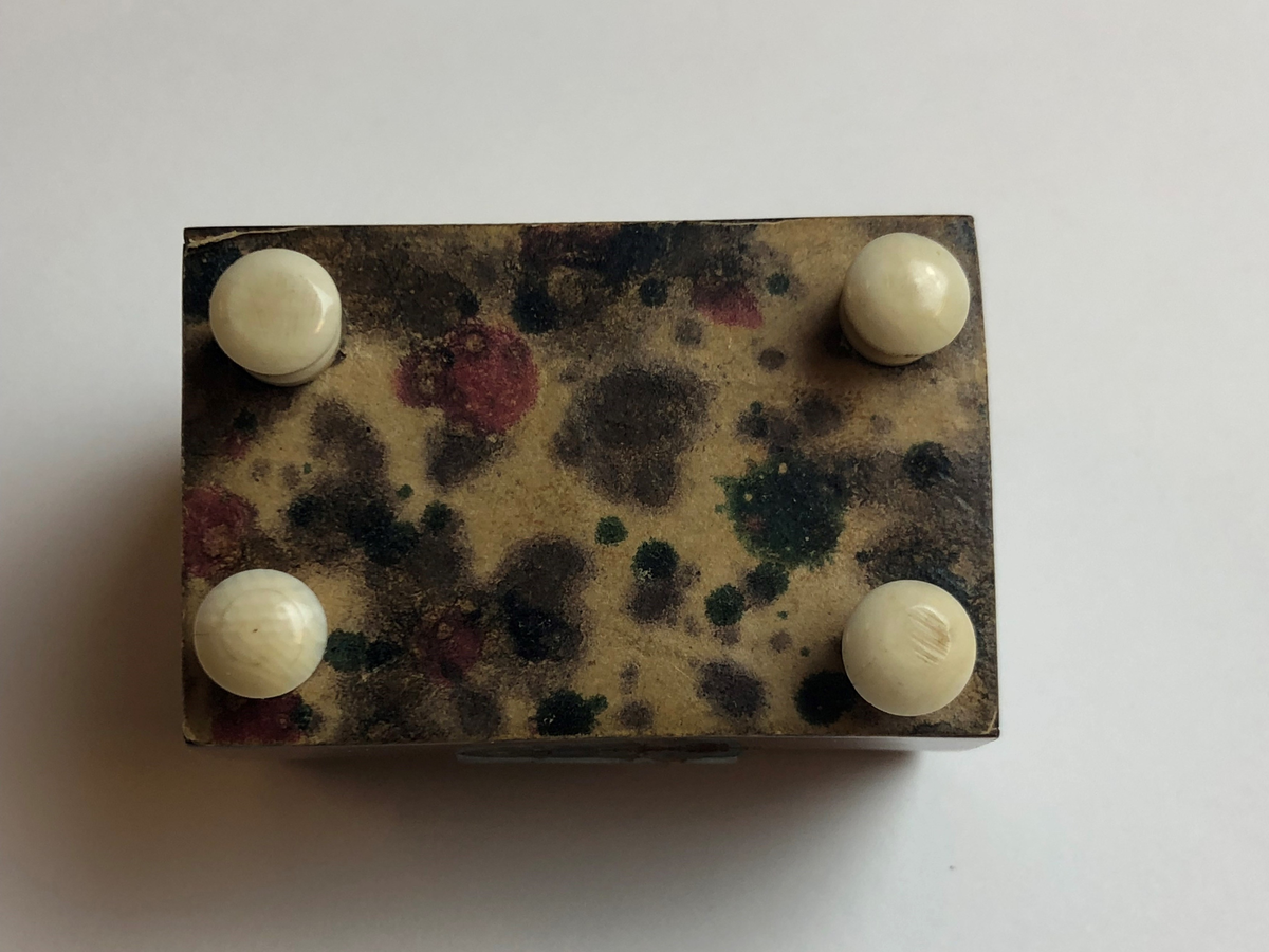Material 4: Papper.
Rektangulär form, underdel med lock som öppnas och stängs med hjälp av ett gångjärn i metall. Av trä,  på utsidan klädd med en yta av ljusbrun plast med mörkbruna fläckar, som ska likna ädelträ. Locket något välvt vilket gör att asken liknar en kista. Underdelens insida klädd med rosa papper, lockets insida klädd med sammet. Lockets ovansida dekorerad med metallpärlor. På undersidan fyra fötter i vit plast.