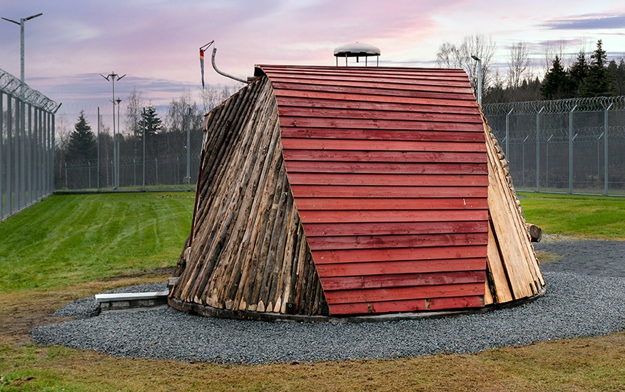 Tekst på nordsamisk lenger ned / Geahca sámegiela teavstta vuolábealde

"Flukthus/Báhtarangoahti" er et kunstverk utført av Vuogas, arkitekturkollektivet med kunstnerne og arkitektene Joar Nango og Eystein Talleraas, i nært samarbeid med innsatte og ansatte ved Ila. Verket har formen og funksjonen til en gamme, men er ikke bygget helt slik en gamme tradisjonelt blir bygget. Hovedstrukturen og de bærende elementene er tilvirket og satt sammen på tradisjonelt vis, men uten jord, torv og never slik det samiske folket gjennom tidene har benyttet seg av. Som vanlig når Vuogas engasjeres i prosjekter, har de undersøkt, funnet og brukt lokale materialer som bærer med seg historikken fra stedet. I dette tilfellet trevirke fra skogen rundt Ila, i tillegg til funnet materiale og gjenstander fra fengselet og gartneriets område. "Flukthus/Báhtarangoahti" er et arkitektonisk kunstverk, men også et oppholdssted og et sosialt rom. Det er bygget for avdelingen, med tanke for de innsatte og ansatte ved NFFA, og der intensjonen har vært at gammen og bålplassen skal bli et godt sted å være.

Joar Nango (f. 1979) er en samisk-norsk kunstner og arkitekt som bor og jobber i Tromsø. Han er utdannet arkitekt fra NTNU i Trondheim og grunnlegger av arkitekturkollektivet Vuogas sammen med Eystein Talleraas (f. 1980). Talleraas bor og jobber i Oslo, og er utdannet arkitekt fra NTNU i Trondheim.

"Báhtarangoahti/Flukthus" lea dáiddadagus, man Vuogas, dáiddára ja arkiteavtta guoktá Joar Nango ja Eystein Talleraas arkitektuvrakollektiiva lea ráhkadan, lagas ovttasbarggus Ila giddagasa fá¿ggaiguin ja virggehasaiguin.  Dáiddadahkosis lea goadi hápmi ja doaibma, muhto dat ii leat huksejuvvon jur dego goahti árbevirolaccat huksejuvvo. Riggi ja válddahagat leat ávdnejuvvon ja cohkkejuvvon árbevirolaš vuogi mielde, muhto goadis eai leat lavnnjit, darffit ja beassit, maid sápmelaccat áiggiid cada leat geavahan godiineaset. Nugo dábálaccat go Vuogas bálkáhuvvo prošeavttaide, de lea arkiteaktakollektiiva guorahallan, gávdnan ja geavahan báikkálaš ávdnasiid, main lea lagasbirrasa historihkka. Dán oktavuodas lea ávnnasmuorat vižžojuvvon vuovddis Ila birra, ja lassin leat gávdnon ávdnasat ja biergasat giddagasas ja gilvvagárdedoalu guovllus. "Báhtarangoahti/Flukthus" lea arkitektuvrralaš dáiddadagus, muhto maiddái orostallansadji ja servvoštallanlatnja. Dat lea huksejuvvon ossodaga várás, NFFA [našuvnnalaš nannejuvvon oktasašossodaga] fá¿ggaid ja virggehasaid gorrái, ja man áigumuš  lea leamaš ahte goahti ja árran galget šaddat sadjin gos lea vuogas leahkit.

Joar Nango (r. 1979) lea Norgga beali sámi dáiddár ja arkiteakta, gii ássá ja bargá Romssas. Son lea oahppan arkiteaktan NTNU:s [Norgga teknihkalaš-luonddudiedalaš universiteahtas] Tråantes/Troanddimis, ja lea vuoddudan Vuogas arkitektuvrakollektiivva ovttas Eystein Talleraas:in (r. 1980). Talleraas ássá ja bargá Osloves, ja lea oahppan arkiteaktan NTNU:s Tråantes/Troanddimis.
