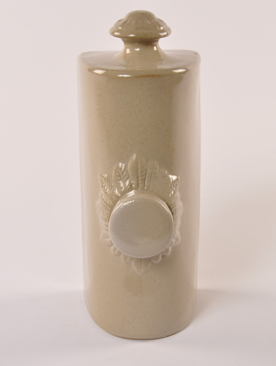 Varmeflaske av gråglassert keramikk, sylinder med flat bunn. Håndtak i ene kortenden og skrukork midt på toppen med enkel bladekor rundt.