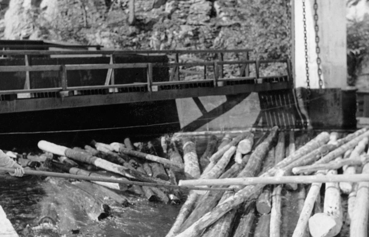 Fra den indre delen påstikkingslensa ved innløpet til tømmerrenna som skulle ta fløtingsvirket forbi kraftverksdammen ved Solbergfoss i Østfold (Nedre Glomma). Påstikkingslensa var traktformet. Den ledet tømmer som drev sakte med strømmen over den store vannflata kraftverksdammen var mot det stedet der renna gikk inn i en forholdsvis trang tunnel i fjellet. Da dette fotografiet ble tatt var tilsiget av tømmer muligens vel stort. I forgrunnen på bildet ser vi (til venstre) ei hand på et hakeskaft. Fløterhakene ble her, når det var nødvendig, brukt til å justere flytretningen til de stokkkene som var på veg inn i renna. Tømmerrenna ved Solbergfoss var cirka 700 meter lang. De øverste 400 meterne gikk gjennom en tunnel som var drevet gjennom berget på den nordvestre sida av kraftverksdammen. Lengre nede gikk renna cirka 300 meter i åpent lende før den endte tilbake i elveløpet. Gjennom tømmerrenna ved Solbergfoss kunne det ekspederes opptil 12 000 tømmerstokker i timen dersom innmatinga ble effektivt utført. Vannet var verdifullt for kraftverket, og fløterne hadde ikke lov til å slippe mer en 15 kubikkmeter per sekund på renna. Vanntilførselen ble regulert via en «inntaksskuff», en hev- og senkbar stålterskel som befant seg under vannspeilet. Posisjonen til denne konstruksjonen kunne justeres etter vekslende vannstand i kraftverksdammen via et kjettingspill fra huset som sto på de kraftige betongsøylene ved innløpet til tømmerrenna.