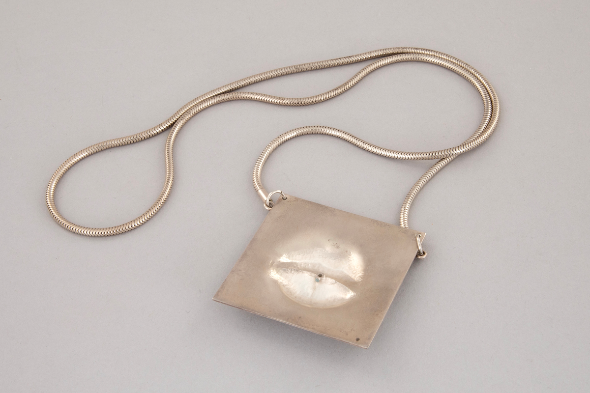 Kvadratisk anheng i sølv med lepper i relieff. I midten av de lett åpnede leppene er det en innfattet diamant. Gravert tekst over: DIAMONDS ARE WOHMENS BEST FRIEND. Et slangekjede i sølv er festet til anhengets øvre hjørner.