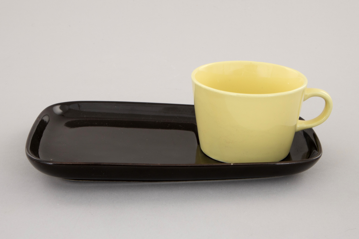 Gul kaffekopp på svart avlangt fat. Fatet er smalere i den enden. Fordypning i fatet for koppen.