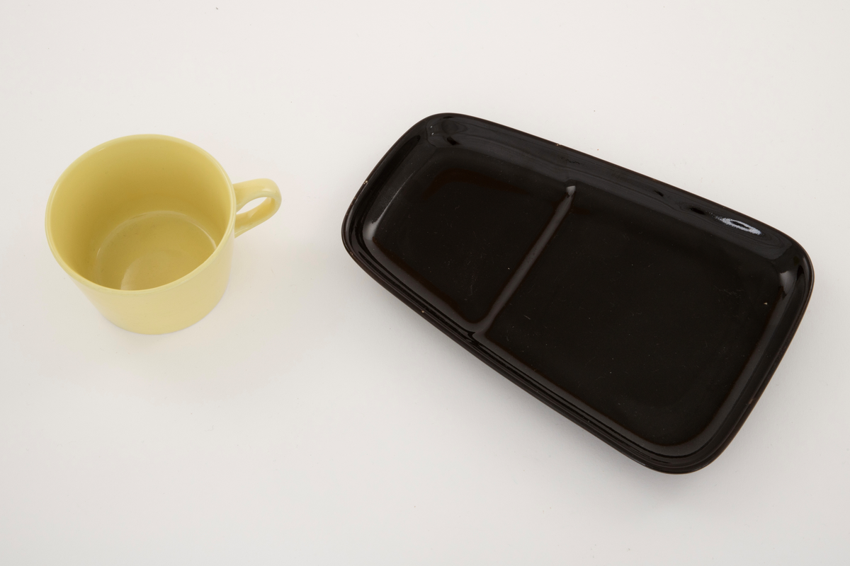 Gul kaffekopp på svart avlangt fat. Fatet er smalere i den enden. Fordypning i fatet for koppen.