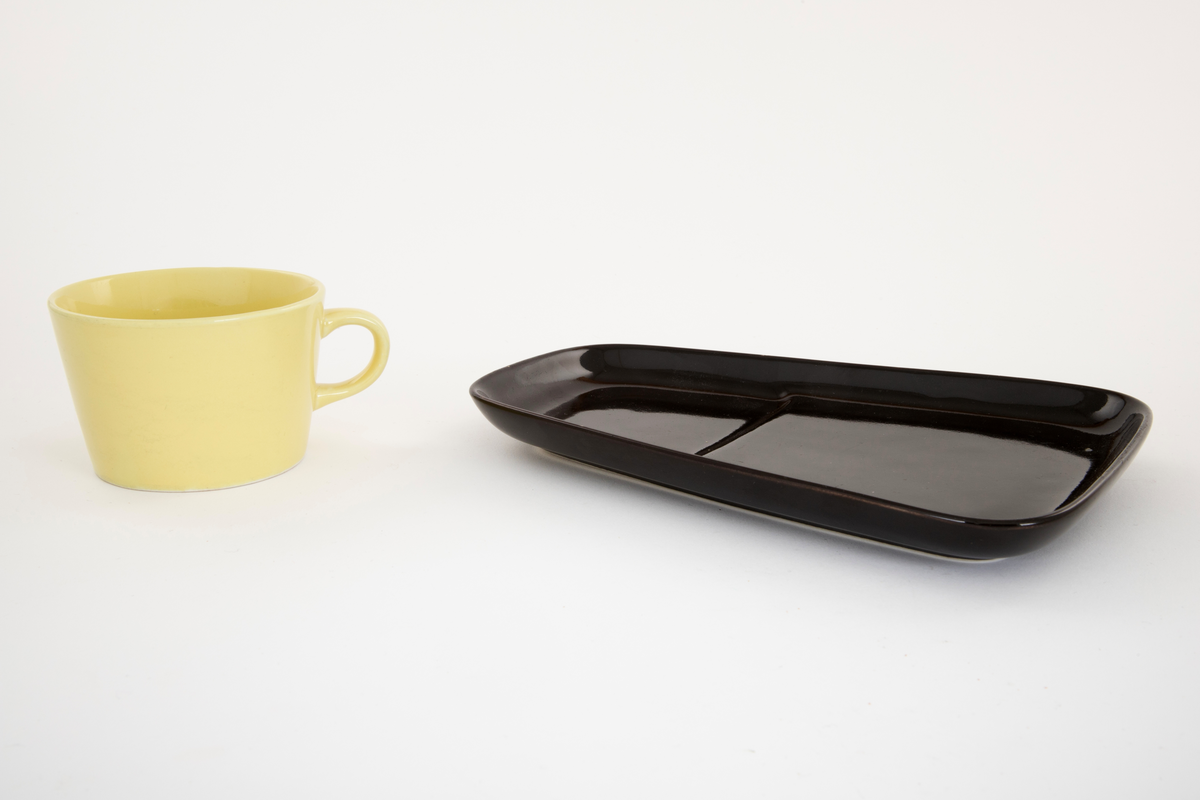 Gul kaffekopp på svart avlangt fat. Fatet er smalere i den enden. Fordypning i fatet for koppen.