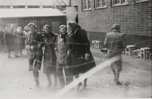 Fotografi tatt inn i kvinnenes luftegård i Grini fangeleir. Til høyre ser man nederste del av hovedbygningen, murbygning, midt på bildet er det fire kvinner som går arm i arm. Man ser piggtrå over bilde.