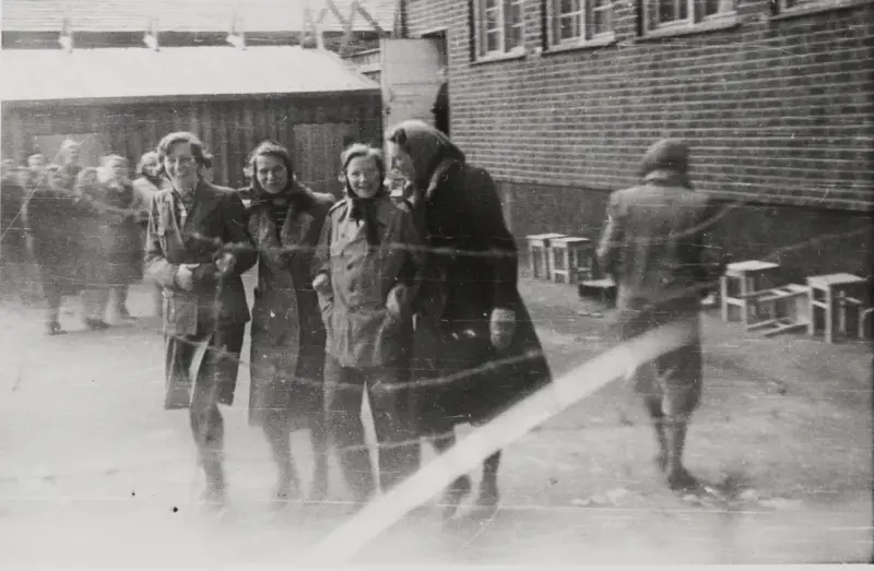 Fotografi tatt inn i kvinnenes luftegård i Grini fangeleir. Til høyre ser man nederste del av hovedbygningen, murbygning, midt på bildet er det fire kvinner som går arm i arm. Man ser piggtrå over bilde.