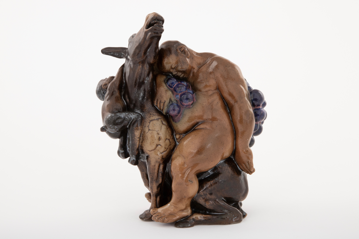 Figurgruppe i stentøy med en stor, sovende mannsfigur, sittende på et esel, drueklaser, og en liten figur som henger i eselets ører. Den lille fiiguren er en faun eller satyr, den store er antagelig en fremstilling av Bacchus. Figurgruppen er brunglasert i lyse og mørke nyanser, samt blått og fiolett.