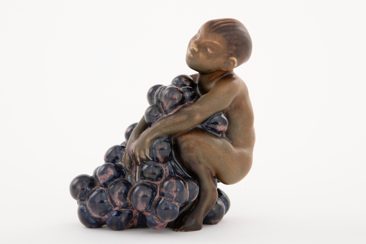 Figur i stentøy; en ung faun med drueklaser. Figuren er brunglasert i lyse og mørke varianter, samt blått og fiolett.
