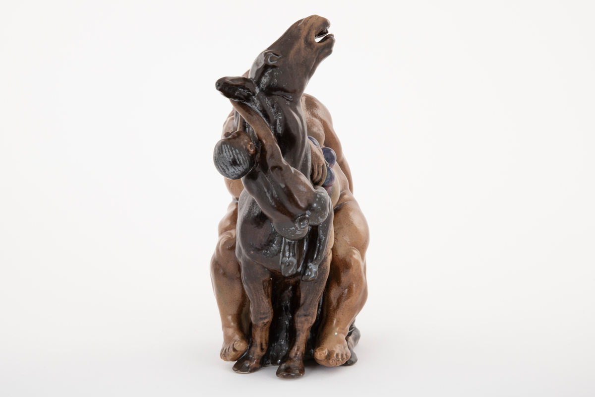 Figurgruppe i stentøy med en stor, sovende mannsfigur, sittende på et esel, drueklaser, og en liten figur som henger i eselets ører. Den lille fiiguren er en faun eller satyr, den store er antagelig en fremstilling av Bacchus. Figurgruppen er brunglasert i lyse og mørke nyanser, samt blått og fiolett.